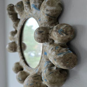 Puede incluir: Espejo redondo decorativo con un marco texturizado único. El marco es de color beige neutro con detalles azules y una serie de formas redondeadas y orgánicas. El espejo está montado en una pared blanca, mostrando su diseño artístico.