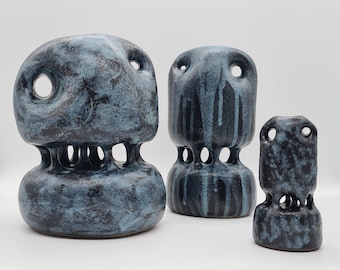 Op de afbeelding: Drie abstracte keramische sculpturen in verschillende maten. De sculpturen zijn donkerblauw met een getextureerde afwerking en hebben grote cirkelvormige openingen en een reeks kleinere gaten. De vormen hebben een unieke, organische vorm.