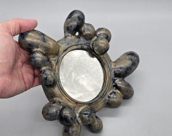 Pode incluir: Um espelho artesanal com um design orgânico incomum. A moldura apresenta um acabamento texturizado e salpicado em tons de bege, azul e preto. O espelho é oval e é mantido pelas extensões bulbosas e irregulares da moldura.