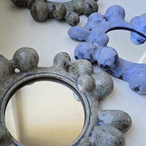 May include: Three decorative mirrors with organic, blob-like frames. The mirrors have a circular shape with a reflective surface. Two mirrors are a muted grey colour, and one is a light blue. The mirrors are placed on a white surface.