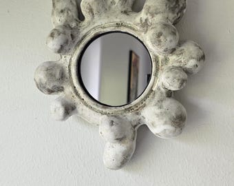 May include: A circular mirror framed by an irregular, organic-shaped border with multiple protruding, bulbous forms. The frame features a textured, off-white finish with mottled grey and dark brown weathering throughout its uneven, bumpy surface.