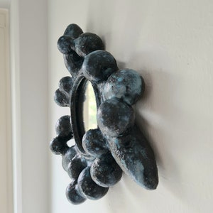 Puede incluir: Un espejo redondo con un marco único y texturizado hecho de cerámica negra y azul. El marco está formado por muchas pequeñas formas redondeadas que le dan un aspecto burbujeante.