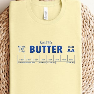 May include: A yellow t-shirt featuring a blue graphic that reads "SALTED BUTTER AA" and includes a measurement guide for tablespoons and cups. The graphic is reminiscent of a butter stick label.