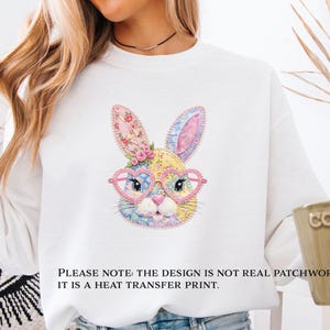 Peut inclure: Sweat-shirt blanc avec un visage de lapin coloré portant des lunettes en forme de cœur. Le motif de lapin comprend des motifs floraux et des couleurs pastel. Le texte sur l'image se lit comme suit : "PLEASE NOTE: THE DESIGN IS NOT REAL PATCHWORK: IT IS A HEAT TRANSFER PRINT."