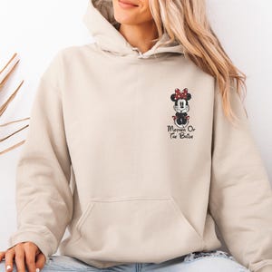 May include: Beige hoodie featuring a Minnie Mouse design with a red bow and the text "Mother Of The Bride" in black. The hoodie has a front pocket and a hood. The design is on the left chest area of the hoodie.