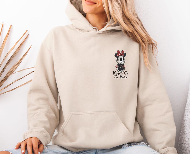 May include: Beige hoodie featuring a Minnie Mouse design with a red bow and the text "Mother Of The Bride" in black. The hoodie has a front pocket and a hood. The design is on the left chest area of the hoodie.