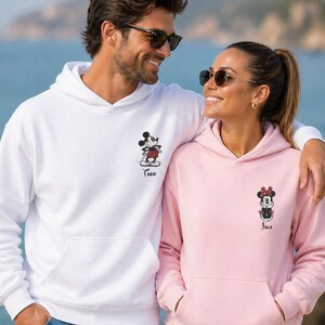 May include: Matching white and pink hoodies featuring Mickey and Minnie Mouse designs. The white hoodie has a Mickey Mouse graphic with the name "Theo" below. The pink hoodie has a Minnie Mouse graphic with the name "Jola" below. Both hoodies have a front pocket.