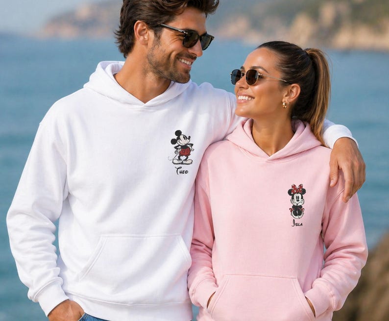 May include: Matching white and pink hoodies featuring Mickey and Minnie Mouse designs. The white hoodie has a Mickey Mouse graphic with the name "Theo" below. The pink hoodie has a Minnie Mouse graphic with the name "Jola" below. Both hoodies have a front pocket.