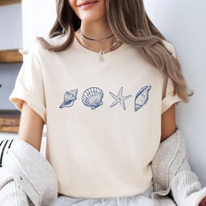 Peut inclure: T-shirt crème avec un motif de coquillages bleus. Le motif comprend un coquillage, une coquille Saint-Jacques, une étoile de mer et un autre coquillage. Le t-shirt a des manches courtes et une coupe décontractée, pour un look décontracté.