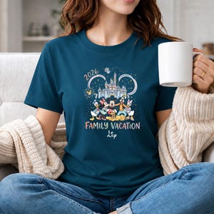 May include: A teal t-shirt featuring a graphic of Disney characters, a castle, and the text "2026 Family Vacation". The design includes Mickey Mouse, Minnie Mouse, Donald Duck, Daisy Duck, and Goofy. The shirt is a crew neck and has short sleeves.