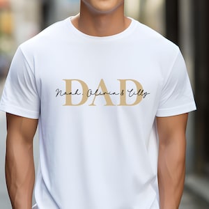May include: White t-shirt with the word "DAD" in gold letters and the text "Noah, Olivia & Tilly" in black script.