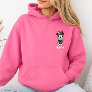 May include: A bright pink hooded sweatshirt featuring a Minnie Mouse graphic with a red bow and the word "Bride" in white. The sweatshirt has a front pocket and a drawstring hood. The design is on the left chest area.