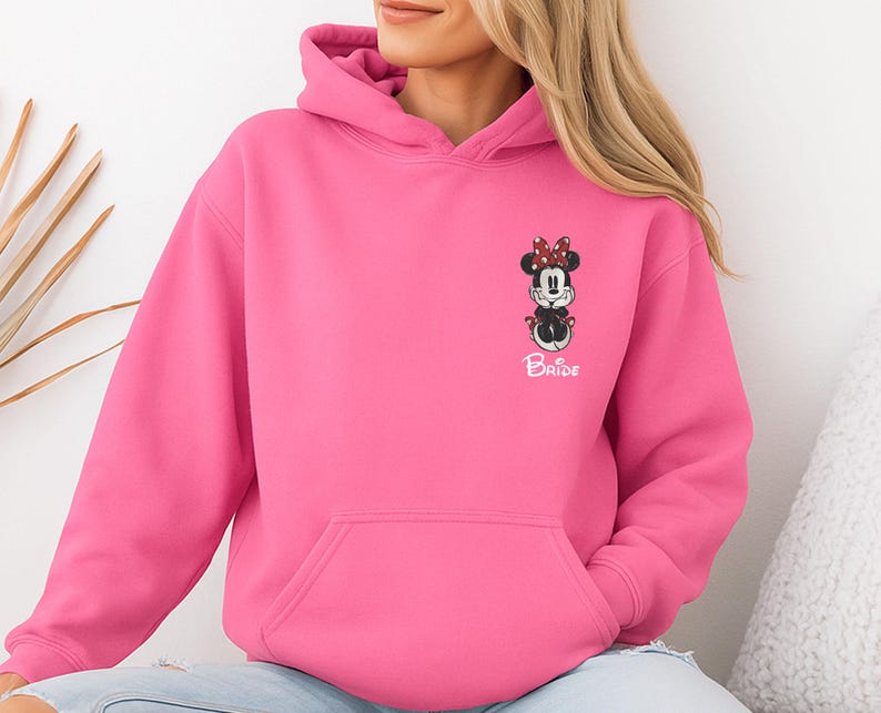 May include: A bright pink hooded sweatshirt featuring a Minnie Mouse graphic with a red bow and the word "Bride" in white. The sweatshirt has a front pocket and a drawstring hood. The design is on the left chest area.