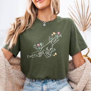 May include: Olive green t-shirt featuring a hand-drawn graphic of two hands holding each other, with the word "Mummy" and "Names" in script. The design includes floral accents in pink, yellow, and blue.