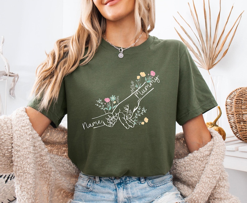 May include: Olive green t-shirt featuring a hand-drawn graphic of two hands holding each other, with the word "Mummy" and "Names" in script. The design includes floral accents in pink, yellow, and blue.