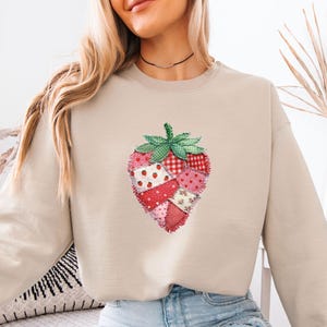 May include: A beige crewneck sweatshirt featuring a large, central graphic of a strawberry constructed from various red, pink, and white fabric patches with frayed edges. The strawberry has a green checkered leaf top and a textured appearance.