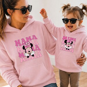 May include: Matching pink hoodies featuring Minnie Mouse graphics. The adult hoodie has "MAMA" stacked text, while the child's hoodie says "MINI". Both hoodies have a front pocket and a drawstring hood.
