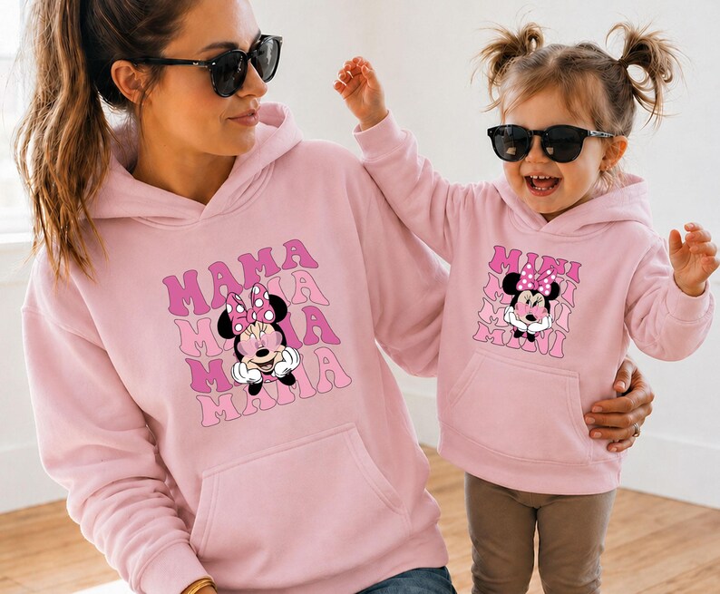 May include: Matching pink hoodies featuring Minnie Mouse graphics. The adult hoodie has "MAMA" stacked text, while the child's hoodie says "MINI". Both hoodies have a front pocket and a drawstring hood.
