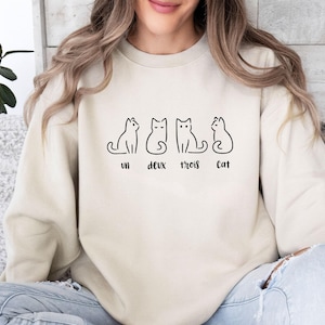 May include: A cream-coloured sweatshirt with a black line drawing of four cats in various poses. The words "un deux trois cat" are printed beneath the cats. The sweatshirt is made of a soft material.