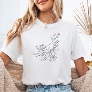 May include: White t-shirt with a black line drawing of hands holding each other, with the word "Nanny" and the word "Name" written on the shirt. The design includes floral accents in pink, yellow, and green.