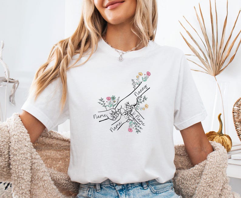 May include: White t-shirt with a black line drawing of hands holding each other, with the word "Nanny" and the word "Name" written on the shirt. The design includes floral accents in pink, yellow, and green.