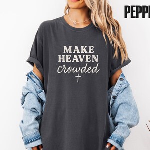Könnte beinhalten: Dunkelgraues T-Shirt mit dem weißen Text "MAKE HEAVEN crowded" und einem Kreuz. Die Ärmel sind hochgekrempelt, und es wird mit einer hellblauen Jeansjacke getragen. Das Wort "PEPPER" wird oben rechts angezeigt.