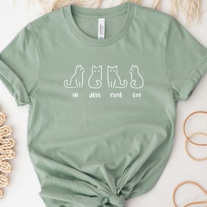 Peut inclure: T-shirt vert sauge avec un dessin blanc de quatre chats et les mots "un deux trois cat". Le t-shirt est en tissu doux et a des manches retrouss&eacute;es. Un design simple et charmant.