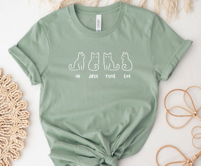 Peut inclure: T-shirt vert sauge avec un dessin blanc de quatre chats et les mots "un deux trois cat". Le t-shirt est en tissu doux et a des manches retrouss&eacute;es. Un design simple et charmant.