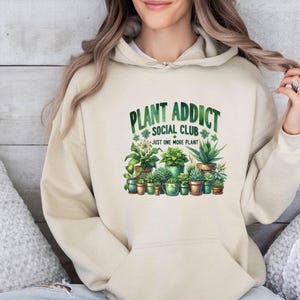 May include: A beige hooded sweatshirt featuring a graphic print of various potted plants and succulents. The text reads "PLANT ADDICT SOCIAL CLUB" in green letters above the plants, with the phrase "JUST ONE MORE PLANT" printed below them.