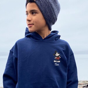 May include: A navy blue hooded sweatshirt featuring a small Mickey Mouse graphic on the front. The cartoon character is depicted in his classic outfit. The name "Theo" is embroidered below the graphic. The person in the image is wearing a dark blue beanie.
