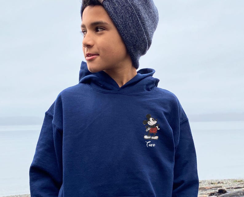 May include: A navy blue hooded sweatshirt featuring a small Mickey Mouse graphic on the front. The cartoon character is depicted in his classic outfit. The name "Theo" is embroidered below the graphic. The person in the image is wearing a dark blue beanie.