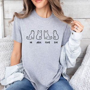 Peut inclure: Un t-shirt gris clair avec quatre dessins de chats noirs stylis&eacute;s. En dessous des chats, les mots "un, deux, trois, Cat". Le t-shirt est fait d'une mati&egrave;re douce.