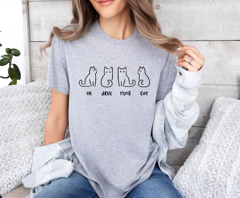 Peut inclure: Un t-shirt gris clair avec quatre dessins de chats noirs stylis&eacute;s. En dessous des chats, les mots "un, deux, trois, Cat". Le t-shirt est fait d'une mati&egrave;re douce.