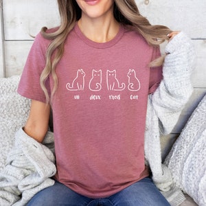 Peut inclure: T-shirt mauve avec un dessin au trait blanc de quatre chats. Le texte "un deux trois cat" est imprim&eacute; en dessous. Le t-shirt est en mati&egrave;re douce, id&eacute;al pour un look d&eacute;contract&eacute;.