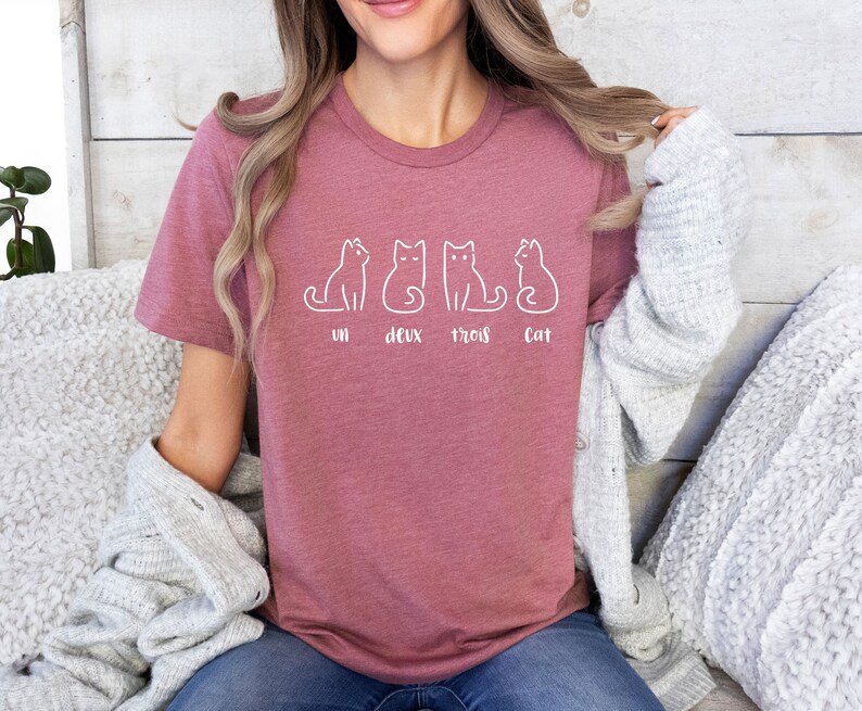 Peut inclure: T-shirt mauve avec un dessin au trait blanc de quatre chats. Le texte "un deux trois cat" est imprim&eacute; en dessous. Le t-shirt est en mati&egrave;re douce, id&eacute;al pour un look d&eacute;contract&eacute;.