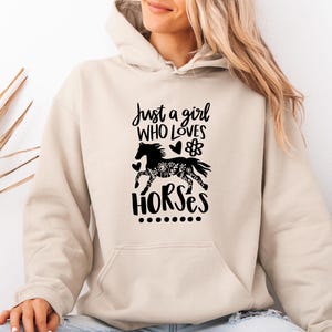 May include: A beige hooded sweatshirt featuring a black graphic print on the front. The text reads Just a girl WHO LOVES HORSES, accompanied by a silhouette of a horse filled with floral patterns, a small heart, a flower, and a row of dots.