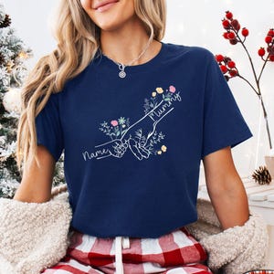 May include: Navy blue t-shirt with a white graphic of two hands holding each other, surrounded by flowers. The word "Mummy" is written above the hands, and the word "Name" is written below. The shirt is a crew neck.