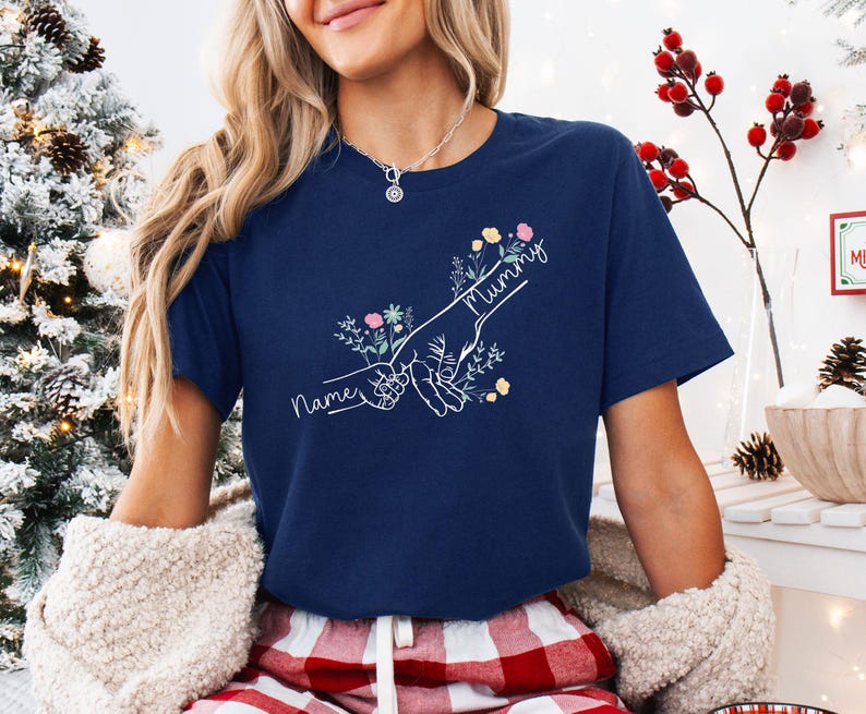 May include: Navy blue t-shirt with a white graphic of two hands holding each other, surrounded by flowers. The word "Mummy" is written above the hands, and the word "Name" is written below. The shirt is a crew neck.