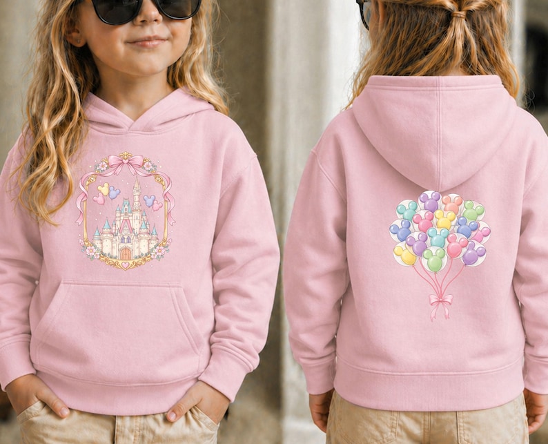 May include: A light pink hooded sweatshirt featuring a front graphic of a castle within a decorative frame, and a back graphic of colorful mouse-shaped balloons. The sweatshirt has a front pocket and a hood.