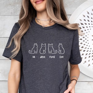 Peut inclure: T-shirt gris fonc&eacute; avec un dessin au trait blanc de quatre chats. Les mots "un deux trois cat" sont imprim&eacute;s en dessous des chats. Le t-shirt est fait d'une mati&egrave;re douce.