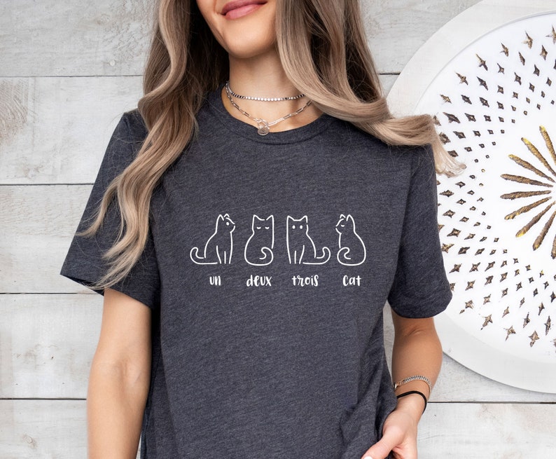 Peut inclure: T-shirt gris fonc&eacute; avec un dessin au trait blanc de quatre chats. Les mots "un deux trois cat" sont imprim&eacute;s en dessous des chats. Le t-shirt est fait d'une mati&egrave;re douce.