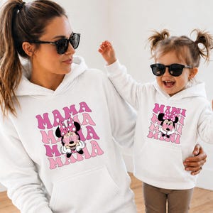 May include: White hoodies with matching Minnie Mouse graphics. One hoodie says "MAMA" in pink retro font, the other says "MINI". Both hoodies feature a cartoon Minnie Mouse with a pink bow. The outfits are worn by a mother and child.