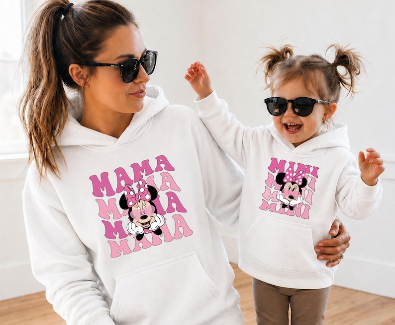 May include: White hoodies with matching Minnie Mouse graphics. One hoodie says "MAMA" in pink retro font, the other says "MINI". Both hoodies feature a cartoon Minnie Mouse with a pink bow. The outfits are worn by a mother and child.