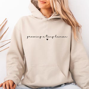 May include: A beige hooded sweatshirt featuring the black cursive text "growing a tiny human" centred on the chest, accompanied by a small black heart icon positioned directly below the text between the words tiny and human.