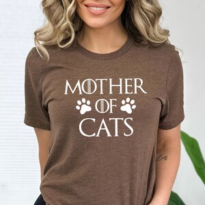 May include: Brown t-shirt with white text that reads "Mother of Cats" with paw prints on either side of the word "of".