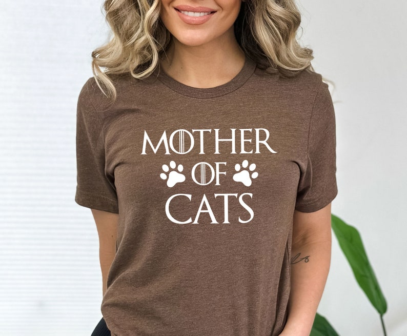 May include: Brown t-shirt with white text that reads "Mother of Cats" with paw prints on either side of the word "of".