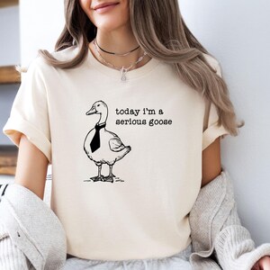 Pu&ograve; includere: Una maglietta bianca con un'illustrazione in bianco e nero di un'oca con una cravatta. Il testo "today i'm a serious goose" &egrave; stampato sulla maglietta.