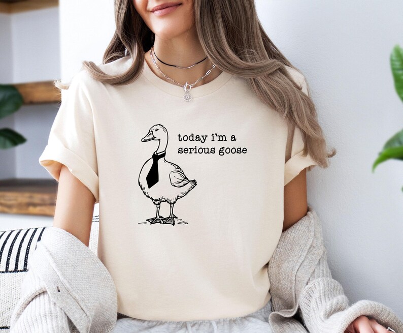 Pu&ograve; includere: Una maglietta bianca con un'illustrazione in bianco e nero di un'oca con una cravatta. Il testo "today i'm a serious goose" &egrave; stampato sulla maglietta.