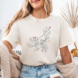 May include: Cream-colored t-shirt featuring a line drawing of hands clasped together, with the word "Mummy" and the names of children. The design includes floral accents in pink, yellow, and green.