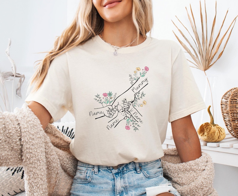 May include: Cream-colored t-shirt featuring a line drawing of hands clasped together, with the word "Mummy" and the names of children. The design includes floral accents in pink, yellow, and green.
